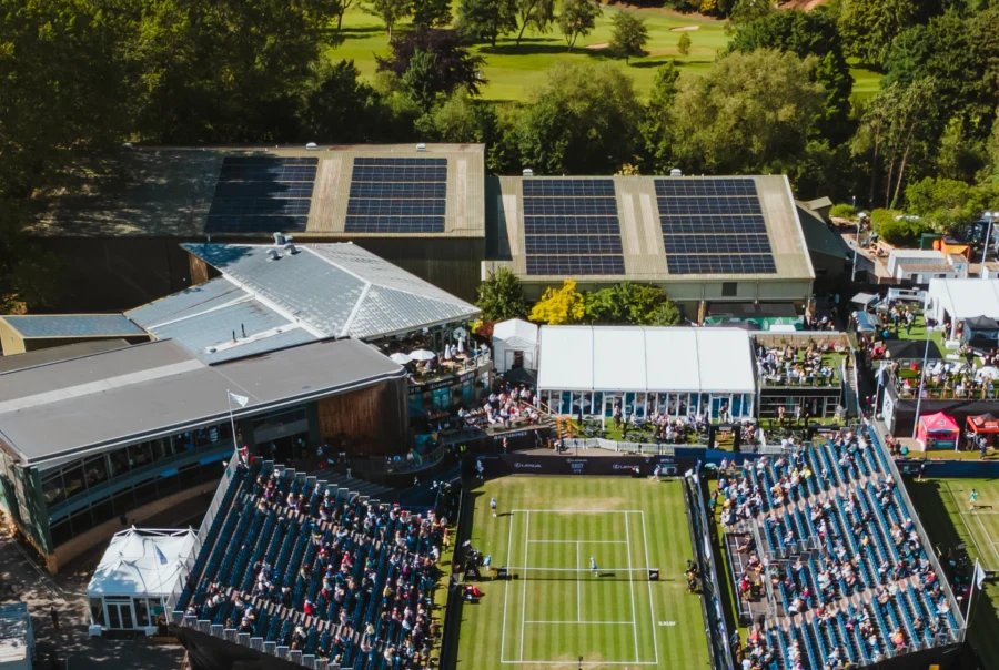 An aerial photo of Ilkley Tennis Club, showing crowds and courts in the foreground, with two roofs covered with solar panels in the top half of the image.
